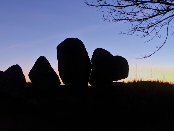 Photo prise au coucher de soleil en hiver : au 1er plan, en haut à droite, la silhouette de quelques branches d'un arbre. Au milieu, la silhouette de 4 grosses pierres de type granitique comme posées l'une à côté de l'autre, qui se découpe sur le fond clair : ciel en dégradé de jaune-orange-bleu.