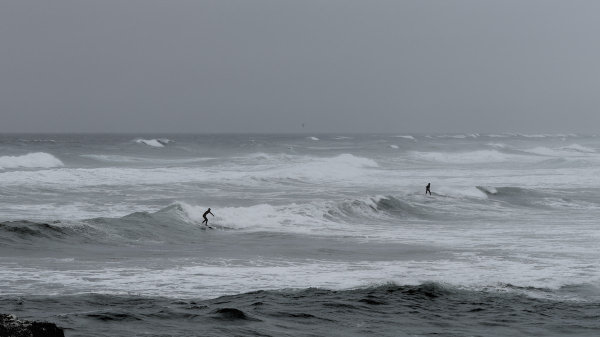 Photo sombre et presque monochrome de grosses vagues sur lesquelles 2 surfeurs essaient de tenir : ils sont en combinaison noire. Les embruns rendent l'atmosphère brumeuse.
Au 1er plan en bas à gauche, on voit un rocher.