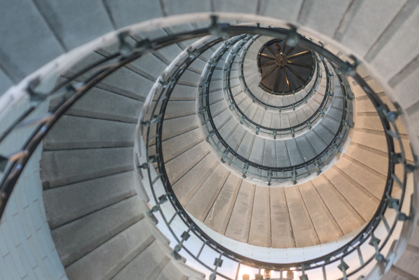 Vue d'en bas d'un escalier en colimaçon dans un phare, les marches sont en pierre et la rambarde en métal. En haut on voit une sorte de couvercle en métal avec des rayons cuivrés.