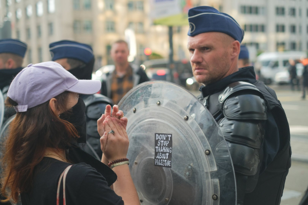 Lors d'une manifestation devant l'ambassade des États-Unis de Bruxelles. Un policier avec un grand bouclier rond et transparent. Le policier a les sourcils froncés et une moue concentré et pas content. sur son bouclier, il y a un sticker colère, un rectangle noir, écrit en blanc "Let's talk about Palestine". Une manifestante se trouve juste devant lui, à quelques centimètres, le visage couvert et elle tape dans ses mains.