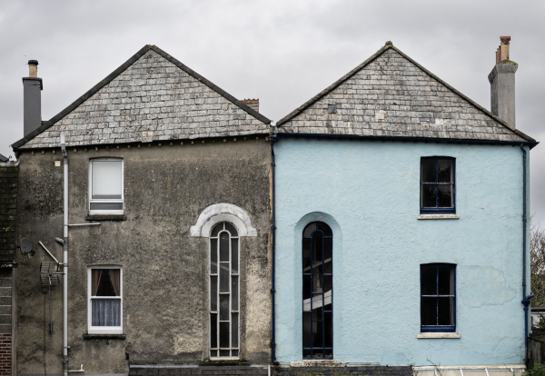 Two houses sharing one wall, unusually each has its own roof. The windows on one are mirror-images of the other. The stairwells are lit by tall windows, side-by-side, in the centre of the photo. The right-hand house is painted a pale blue, the other has plain render.