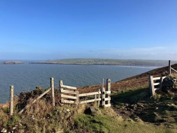 Photo of an open gateway on a coastal footpath, a wooden style adjacent on the left. Beyond it is a view of a bay with a grass covered coastline on the opposite side scattered with white houses. A small island sits off the coast to the left. To the right, waves break on a wide sandy beach at the mouth of a river. The sky is an almost cloudless blue, a haze on the horizon inland.