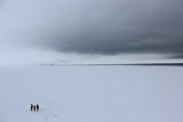 Untere Hälfte große Fläche weißes Seeeis in der Arktis, ein dünner Streifen dunkelblaues Meer trennt das Eis vom wolkenverhangenen, düsteren Himmel. In der unteren rechten Ecke drei Forscherinnen in neon-warnfarbenen Winteranzügen.