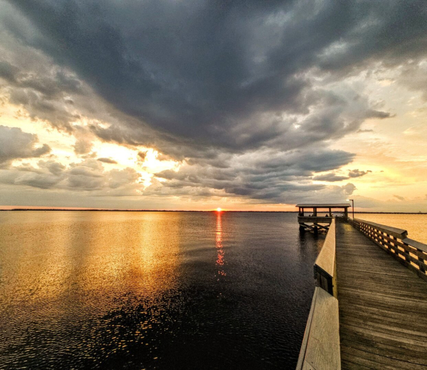 Enormous dark clouds rise from the horizon in the distance in a tornado shaped funnel just above the red hot sun, about to cross below the horizon over a vast, dark river. Each side of the dark funnel cloud formation is brightly illuminated in golden shades from the sun's intensity, which reflects onto the water's surface below.  The red hit sun also casts a red, laser like beam of light across the water's surface as well.  To the right, a large wooden pier with a covered observation deck reaches far out into the river waters, where tiny silhouettes of people settled in to watch the sunset appear within the golden glow cast upon the pier's wooden surfaces.