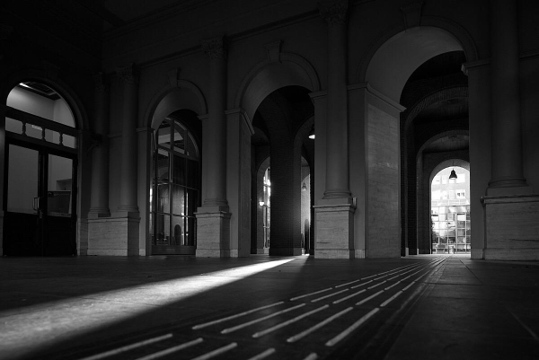 A monochromatic interior image featuring arches and columns. A beam of light highlights the floor, which has a patterned design, with reflections visible in glass doors and windows. The overall atmosphere is serene and architectural, suggesting a spacious foyer or hall.