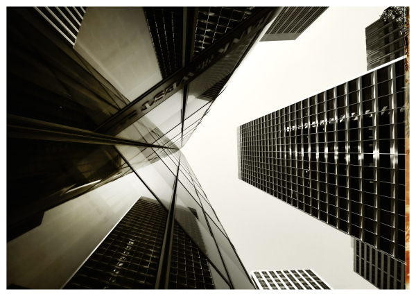 A monochrome view looking up from the ground at a handful of tall buildings along Market Street in downtown San Francisco. 

On the left is the Shacklee building... a modern building with folds and large panes of mirrored glass. Here, the interior angle of one of those folds has reflections of the tallest building directly across the street.

Directly overhead, the sky is gray and glaring. A marine layer of clouds (fog) has covered the city.