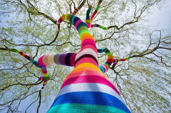 A photo of a weeping willow tree, photographed upwards from its base. The trunk and branches are encased in multi colored knitting, with stripes reaching up many meters into the branches while we still stand under an unbrella of green leaves from the tree too