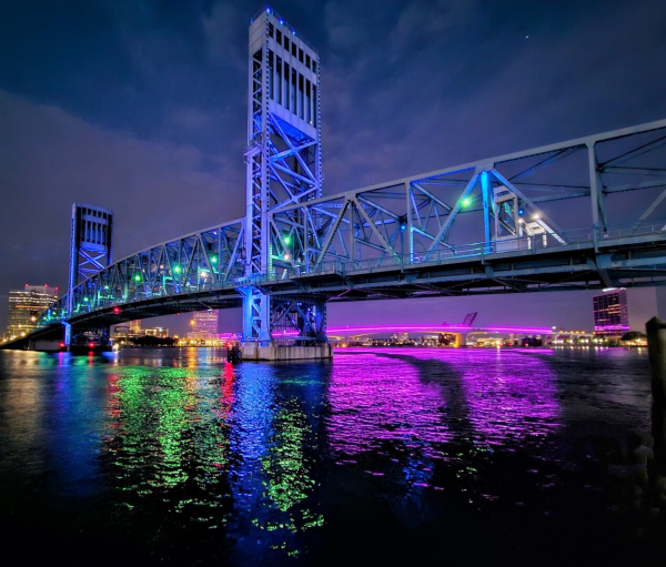 From the northbank of the Saint Johns River, downtown Jacksonville, a late night view across the river where two of Jacksonville's many bridges are visible with bright colorful illumination, which along with the city skyline,  and late night traffic, add a rainbow of colorful reflections across the river's dark surface.