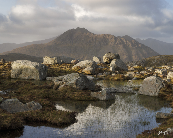 a small lochan high in the remote mountains of scotland reflects beautiful clouds and blue skies. erratic boulders are strewn across the foreground while the background features a shapely mountain with a steep and wandering ridgeline. soft evening light dances across the scene. 