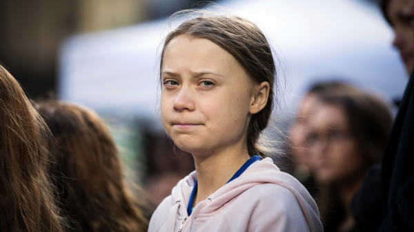 Greta Thunberg, wearing a blue t-shirt and a pink hoodie, looking at the camera during a meeting, seems to feel angry or unsatisfied.