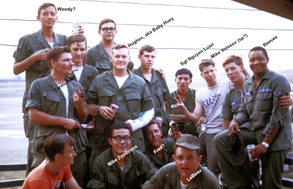 Thirteen US GIs plus one Vietnamese Air Force sergeant standing on balcony. Most dressed in Vietnam era fatigues, some holding cans of beer with Danang Air Base in background. Circa 1969.