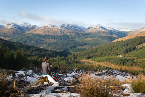 woman sitting on fallen trees with snow, looking at distant mountains