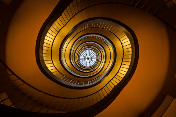 Look up view of spiral stairs in Paris with orange walls and golden electric lighting, with a  round skylight in the center at the top middle showing the blue sky and looking like an eye. 