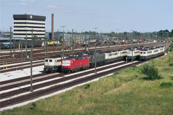 Das Foto zeigt den Rangierbahnhof Maschen, einen der größten Rangierbahnhöfe Europas, an einem sonnigen Tag. Im Vordergrund stehen mehrere Elektrolokomotiven verschiedener Baureihen auf parallel verlaufenden Gleisen. Links befindet sich eine beige-blaue Lok der Baureihe 140, direkt daneben eine moderne, rot lackierte Lok der Baureihe 152. Dahinter sind eine grüne E-Lok der Baureihe 151 sowie mehrere weitere Loks der Baureihen 140 und 110 in der klassischen beige-blauen Farbgebung zu sehen.

Die Gleisanlagen erstrecken sich weit in die Tiefe des Bildes und sind von zahlreichen Oberleitungsmasten gesäumt. In der Ferne erkennt man weitere Loks, darunter rote Exemplare der Baureihen 143 und 145, die auf Abstellgleisen stehen.

Im linken Hintergrund dominiert ein großes, sechsstöckiges Verwaltungsgebäude mit dunkler Fassade das Bild, auf dessen Dach das Logo der Deutschen Bahn (DB) prangt. Daneben befindet sich ein langes, flaches Gebäude, vermutlich eine Werkstatt oder ein Betriebsgebäude. Ein hoher Industrieschornstein ragt über die Anlage hinaus.

Rechts neben den Gleisen ist eine grüne Wiese mit niedrigen Sträuchern und Gräsern zu sehen, die einen Kontrast zur technischen Infrastruktur des Bahnhofs bildet. Die Oberleitungen und Signale sind über das gesamte Gelände verteilt, und die helle Sommersonne wirft deutliche Schatten auf die Bahnanlagen.