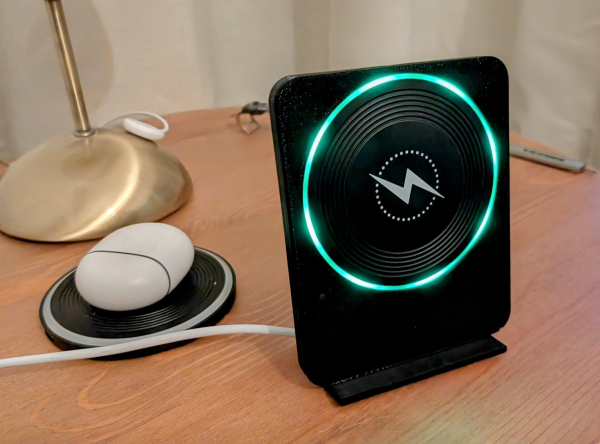 The Qi Charger Stand (activated, because a Pixel 8 pro was 3 seconds before near the charger) with a green light ring stands on a wooden table. Near the stand an unplugged second charger with Pixel Buds pro on top). In the background a lamp Anna Pixel Watch 3 charger).