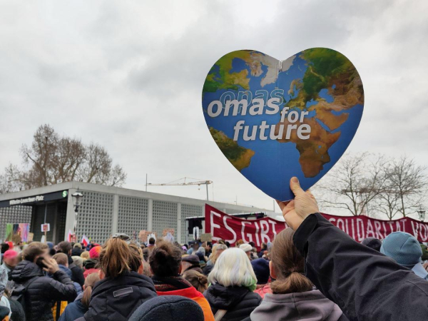 Auf dem Bild ist eine Menschenmenge auf der Kundgebung am kleinen Wilhelm-Leuschner-Platz in Leipzig zu sehen. Eine Hand hält das Logo der Omas for Future - das Erdherz  - in die Höhe.
