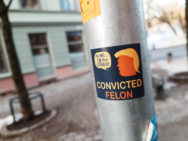 A photograph of a sticker on a lamppost. The sticker features a silhouette of Donald Trump in the top right with a speech bubble which reads "It's me. I'm the loser!" and the text below "Convicted felon"