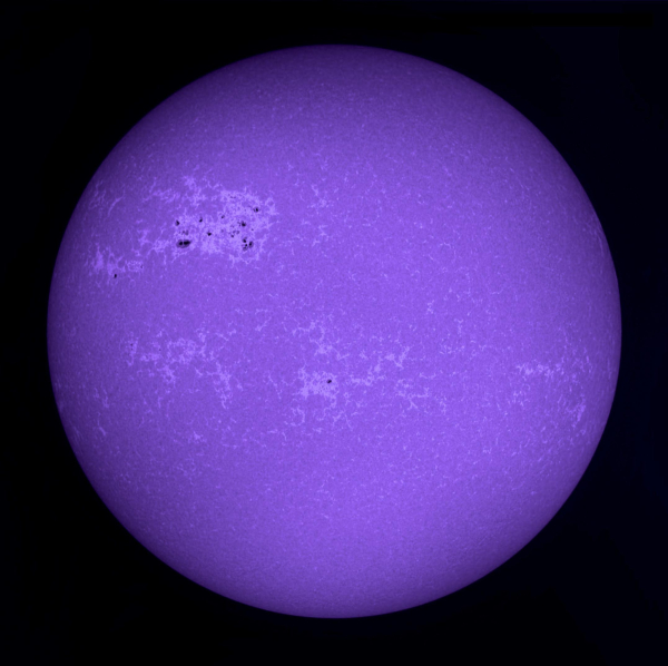 Die Sonne im Licht der Calcium-K-Linie, blauviolett eingefärbt. Eine Ansammlung von Sonnenflecken oben links ist von ausgedehnten hellen Bereichen umgeben, weitere ausgedehnte helle Bereiche umgeben einen kleinen Fleck unterhalb der Bildmitte 