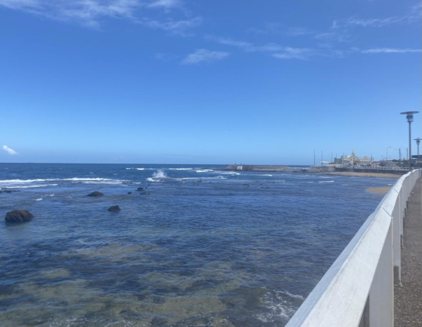 Looking south-ish across the Cowrie Hole towards the Newcastle Ocean Baths. The water covers most of the rocks and is clear enough to show the sandy bits too. A few small waves breaking on the edge of submerged rock platform. Blue sky with a few wispy white clouds high up. 