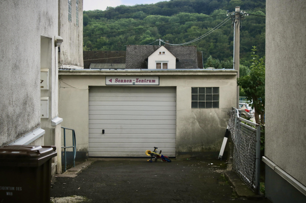 das bild im querformat zeigt eine  dreckige und hell gestrichene garage zwischen zwei hauswänden. über dem rolltor der garage ist ein  weißes schild mit magentafarbener schrift und dem text "sonnen-zentrum". es zeigt mit einem dreieckigen pfeil nach links. auf der linken seite ist offenbar ein durchgang.

die garage ist das zentrale und mittig positionierte element des bildes. davor ist eine einfahrt zu sehen. ein kinderfahrrad liegt vor der garage. das rolltor ist weiß und rechts daneben ist ein fenster mit 4x3 glasbausteinen.
links unten im bild steht an der wand eine schwarze mülltone mit braunem deckel.
die wände der häuser links und rechts der garage sind weiß bzw. hellbeige gestrichen. sie haben deutlich patina über die jahre angelegt.
über der garage wird der blick freigegeben auf eine bewaldete anhöhe. davor sind noch dächer anderer häuser erkennbar.