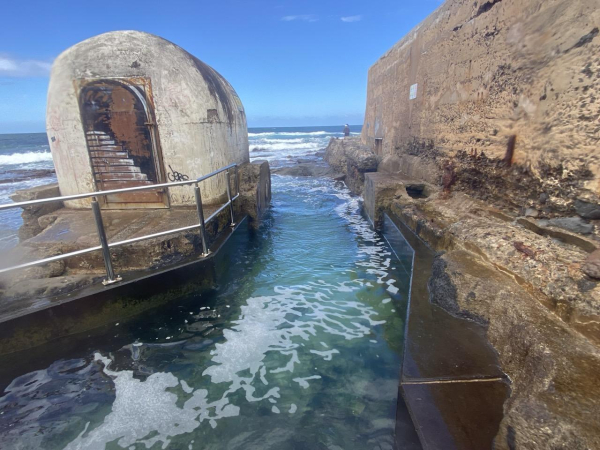 A narrow channel of green sea water (over 1 metre, not quite 2) runs between the rounded white pump house building on the left and the high weathered concrete wall on the right (around 3 metres maybe). The kids (mostly look like teens) jump off the wall into that water. Three of them had just climbed out before i took this one. Ocean horizon in the distance.