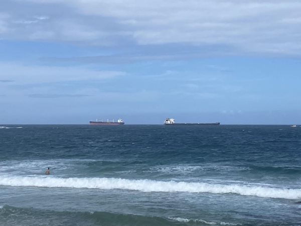 Two big ships, bulk carriers most likely, on the ocean horizon off Nobby's Beach. One is heading left to the Harbour. The other has just left the harbour, heading out to its next port. Blue-green water, blue sky with a light layer of white cloud. Waves breaking in the foreground with one guy about waist deep in the water. Beach just out of view.