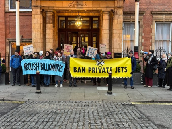 Picture of a group of protesters from Climate Resistance holding placards reading EAT THE RICH, BILLIONAIRES SHOULD NOT EXIST, THEY PROFIT WE BURN,  and two large banners: one blue, reading ABOLISH BILLIONAIRES, and a second yellow, reading BAN PRIVATE JETS