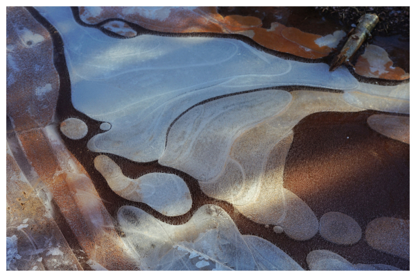 Farbfoto mit weißem Rand. Blick auf eine große vereiste Pfütze, darin Muster aus weißem, bräunlichem und rotem Eis. Es sieht aus wie marmoriert oder wie eine stilisierte Geländekarte.