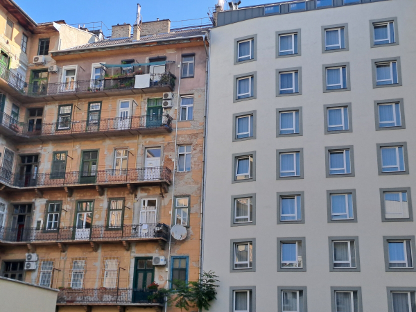 Photo of two apartment buildings that share a wall. The one on the left is an older building with 6 floors, peeling peach colored wall paint, wrought iron balconies, and brick chimneys. The door and window frames are various shapes and colors, and there are various objects on the balconies. The building on the right is new, 8 floors (despite being the same height), with a clean white wall and small, uniform grey windows.