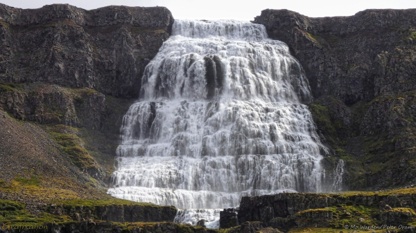 A colour photo of a mountainside under a cloudy sky, with sunlight from the right. In the centre of the shot is a waterfall, dropping from near the top of the frame to almost the bottom. The rock around it is brown and grey, and has vegetation on the more horizontal surfaces. The waterfall itself is white right from the top, as the rockface has steps in it which the water is hitting all the way down. It spreads out into a wedge shape, almost a trapezoid. The top of another falls is visible below it.