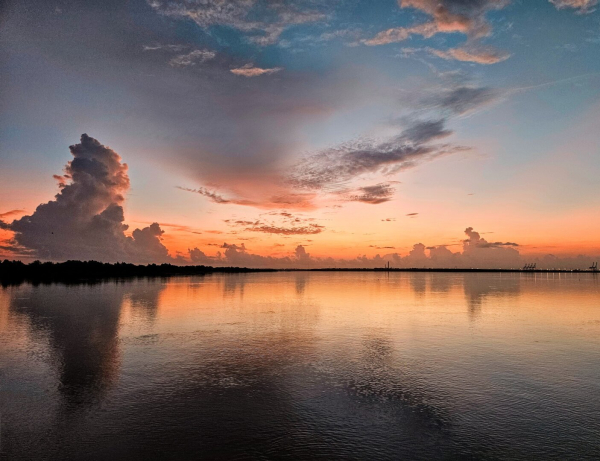 Subdued sunrise over Saint Johns River. Giant cloud formations tower over the landscape, turning enormous sea port cranes into miniature silhouettes along the shoreline of a vast, wide river. Higher above, against a blue sky smaller clouds take a V-shaped pattern, pointing downwards at the shoreline where golden shades rise above the horizon as a sunrise approaches.  All producing reflections upon the dark, still river below.