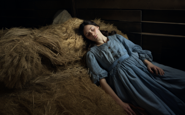 A young woman wearing a 19th century prison dress sleeps on hay bales in a barn loft