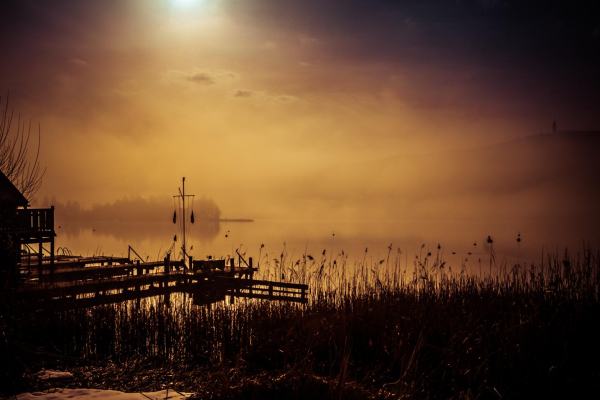 Die Silhouette einer Fischerhütte dahinter der See im Nebel . Schemenhaft erkennt man im Hintergrund das andere Ufer