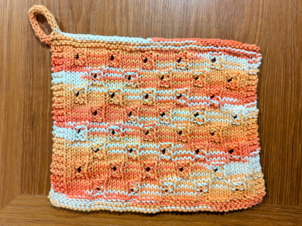 A photo of a knitted cotton dishcloth laid flat on a wooden table. The dishcloth features a basket weave pattern with little eyelet holes in the centre of some of the alternating weave cells. It's in a pleasing orange, yellow, and white variegated colour and has a little loop knit on one corner for hanging it.