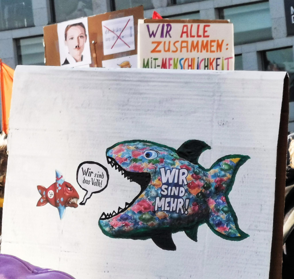 Ein kleiner brauner AfD-Fisch mit der Sprechblase "Wir sind das Volk!" wird von einem großen bunten Fisch mit der Aufschrift "Wir sind mehr!" gefressen.