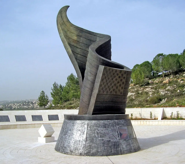 The image depicts a sculptural monument. It features a large, abstract design that resembles a twisted or flowing form, possibly made of metal. The base has inscriptions, and the overall structure seems to be designed to be visually striking, set against a backdrop of trees and a clear sky. This type of monument could be intended to commemorate a specific event or honor individuals or groups.

Image Credits: Wikimedia / dr. avishai teicher User:Avi1111 / Public domain