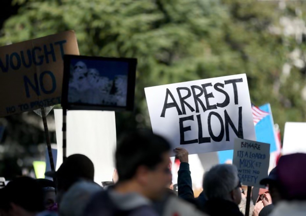 The image shows a crowded protest scene with people holding various signs. A prominently displayed sign reads "ARREST ELON," while other signs reflect different messages. The background features trees and flags, indicating an outdoor setting.