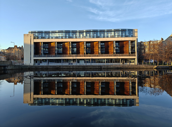 A colour photograph in landscape orientation of a building beside the Water of Leith. The building is of a modern design, comprising three storeys. The river is flat and reflecting the building. The sky behind the building is a gentle blue. The afternoon light bathes the picture in a golden light.