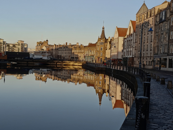 A colour photograph in landscape orientation of the view along the Shore, a road in Leith beside the Water of Leith. 

The river and road curve from the bottom right to the middle left. 

The buildings along the road are bathed in a gentle winter's light, except this nearest the camera which are in shadow. 

The buildings and the deep blue sky are reflected in the still water.