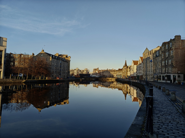 A colour photograph in landscape orientation of the view along the Shore, a road in Leith beside the Water of Leith. 

The river and road curve from the bottom right to the middle left. 

The buildings along the road are bathed in a gentle winter's light, except this nearest the camera which are in shadow. 

The buildings and the deep blue sky are reflected in the still water.

Across the river are former warehouses, deep in shadow which extends to the river nearest the camera.