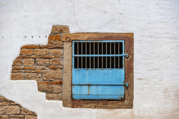 In einem Sandsteingewände ist ein metallener Fensterladen dessen unterer Teil geschlossen ist und die oberer Hälfte ausgeführt mit Gitterstäben. Die Wand ist zum Teil verputzt und an manchen Stellen erkennt man das Sandsteinmauerwerk.

In a sandstone wall there is a metal shutter, the lower part of which is closed and the upper half is made with bars. The wall is partly plastered and in some places the sandstone masonry can be recognised.