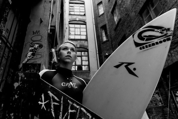 The photograph features a portrait of a female surfer, captured in a vertical composition, positioned against a textured, graffiti-covered wall in an urban alley. She is centrally framed, with her head tilted slightly upward, conveying a sense of aspiration. The surfboard she holds is partially visible on the right side, featuring brand logos. The image is monochromatic, emphasizing light and shadow, and the soft focus on the background draws attention to her facial expression.