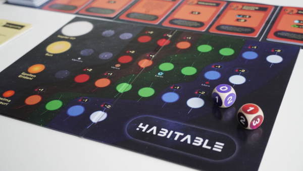 Das Bild zeigt ein Tischspielbrett mit dem Titel „Habitable“, das ein farbenfrohes Layout mit Planeten und Symbolen enthält, die verschiedene Himmelskörper darstellen. Das Spielbrett enthält markierte Spielstände und verschiedenfarbige Kreise. Im Vordergrund befinden sich zwei mehrflächige Würfel, auf deren Flächen Zahlen abgebildet sind. Im oberen Teil des Spielbretts befinden sich Karten mit zusätzlichen Spielelementen.
 („Habitable“ will zwischen den Generationen vermitteln Foto: Heidelberger Instituts für Theoretische Studien (HITS) )