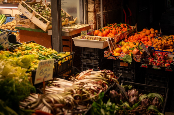 A vibrant market scene featuring an array of fresh fruits and vegetables. Stalls display oranges, strawberries, artichokes, zucchinis, and lettuce, with price tags visible. The setting is dimly lit, creating an inviting atmosphere.