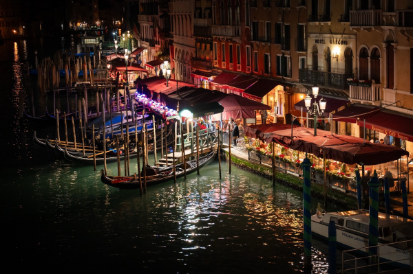 A nighttime scene of a canal in Venice, featuring several gondolas moored alongside colorful restaurant awnings. The water reflects lights from buildings and lampposts, creating a lively atmosphere.