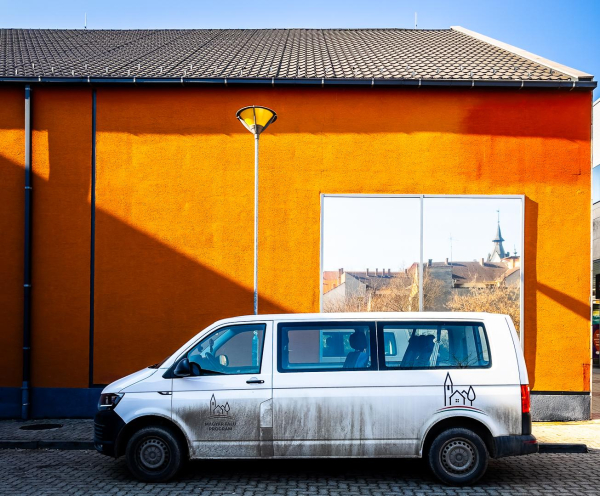 Terrakottafarbene Wand mit einem Fenster, in dem sich gegenüberliegende Häuser spiegeln. Schräger, harter Schatten und ein weißer Kleinbus, der passgenau davor parkt.