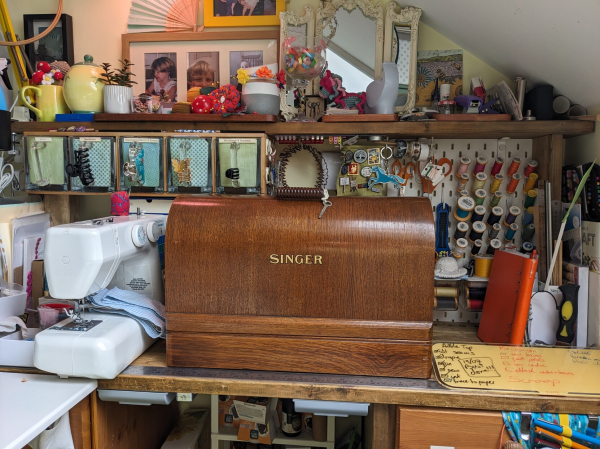 A gleaming wooden bentwood case for a Singer sewing machine, sitting on a very crowded desk, with a plastic Janome sewing machine on its left. Scissors and thread are on a pegboard on the back right.