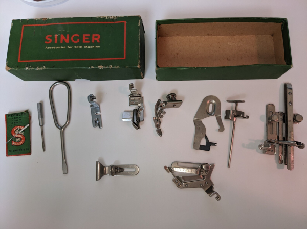 A Singer branded cardboard box, open, behind an array of sewing machine accessories including needles, screwdrivers, presser feet, and unidentifiable twisty bits of stuff