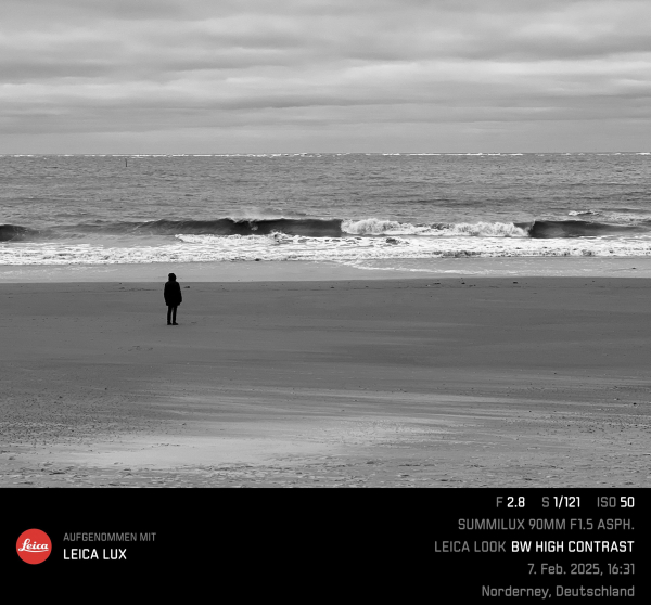 Schwarz-weiß Bild mit hohen Kontrast, ein Mensch an einem ansonsten leeren Strand, den Blick aufs Meer gerichtet, wo eine Welle gerade zu brechen beginnt