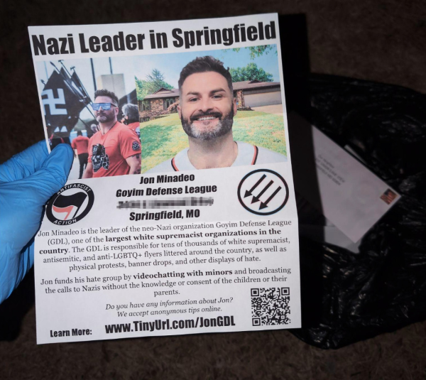 a hand wearing a glove holding a flyer titled "Nazi Leader in Springfield" that is warning that the leader of the neo-Nazi group Goyim Defense League is now living in Springfield, MO. There is a garbage bag on the group full of stamped envelopes in the background.